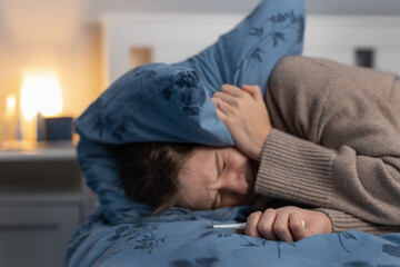 Young caucasian woman suffering from severe headache or migraine. Female suffers from cold and flu, have heat temperature. Seasonal illness. Selective focus on hand with thermometer. Girl feeling sick