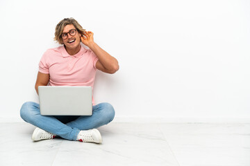 Young caucasian man sitting on the floor with his laptop isolated on white background listening to something by putting hand on the ear