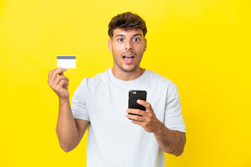 Young caucasian handsome man isolated on yellow background buying with the mobile and holding a credit card with surprised expression
