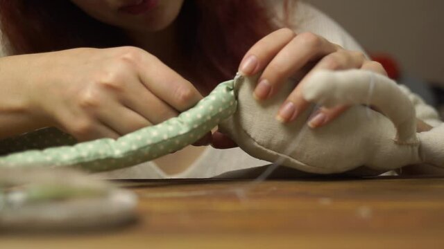 Red-haired girl sews rag doll on wooden table. Handmade toy made of linen, sintepon and synthetics. Process of sewing legs to body of doll. Craft diy.