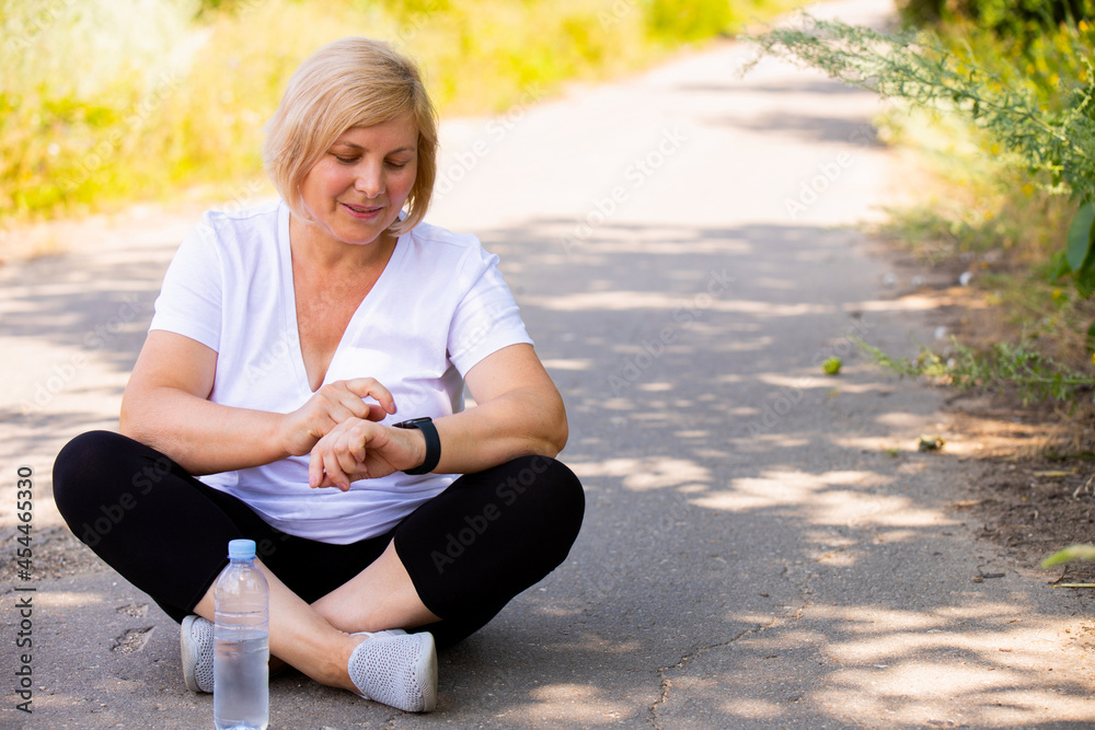 Wall mural The photo of a happy blonde sporty woman sitting in a lotus position on the road in the park wearing sports clothes pointing on the digital watch on her hand.