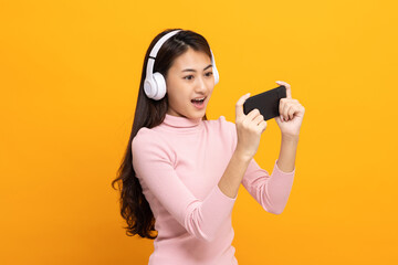 Obraz premium Beautiful young asian women play mobile game and put on wireless headphone standing on isolated yellow background. Playing game on smartphone winning victory moment. Very enjoy and fun relax time