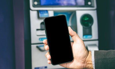 Young businessman standing front of atm machine using smartphone withdraw the money without atm credit card. He ut the pin code into smartphone and take the money.