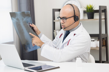 Naklejka na ściany i meble Doctor talks to patient via online video conferencing with computer wearing headset. Explain about taking medications to treat illnesses holding x-ray film.