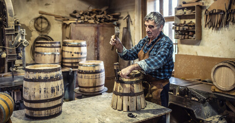 Aged man crafts a new vintage-looking wooden barrel using hammer to put the metal ring around the pieces of wood