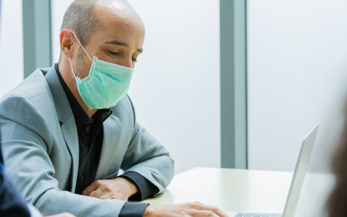 Smart Caucasian businessman wearing formal suit, face mask protect virus, new normal, social distancing, sitting, typing in laptop, using internet and working with intention in indoor office company.