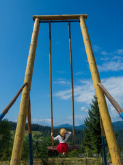 Back of girl in a straw hat, white blouse, red skirt and yellow sneakers sitting on a swing with the mountain view. Calm and quiet wanderlust concept moment when person feels happiness and life.