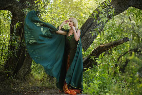 A blonde woman in a green druid cloak and in a fantasy dress with leaves. A sorceress girl in medieval clothes stands in the forest near a reservoir.