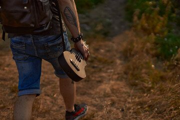 a hippie guy in denim shorts and with a tattoo on his arm walking near the forest, holding a...