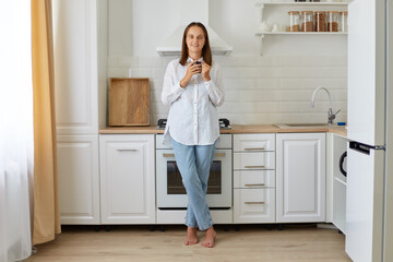 Full length portrait of a calm woman holding a cup of tea or coffee in her kitchen, attractive female wearing white shirt and jeans enjoying hot beverage at breakfast.
