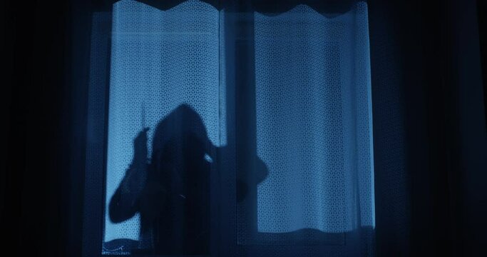 Killer is Looking through the Window at Night Holding a Knife Preparing to Break in the House in Silhouette
