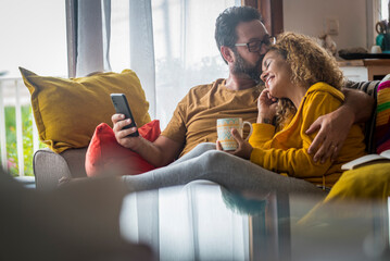 Tenderness time moment for mature caucasian couple at home sitting on the couch together - love and relationship adult people indoor lifestyle - man kissing woman with phone on hand - Powered by Adobe