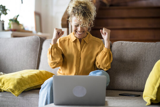 Happy woman celebrate in and exult in front a laptop computer sitting on the sofa at home - concept of win and online work or shopping lifestyle - happy people smart working office job