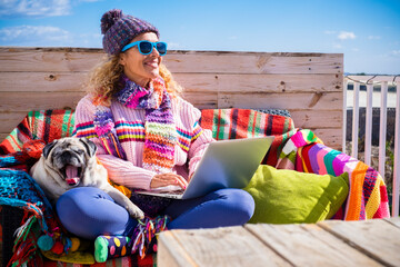 Woman sitting outdoor with dog smiling and enjoying best friend - happy modern people working outside with laptop computer and internet connection - winter clothes and colorful style