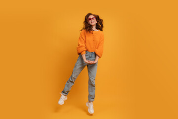 Fototapeta premium Laughing stylish red head woman in orange sweater and blue jeans jumping over yellow background. Full lenght.