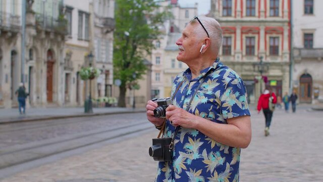 Happy senior tourist with retro photo camera listening to music on earphones, dancing outdoors, having fun alone on city street. Elderly mature and modern devices concept. Active life after retirement