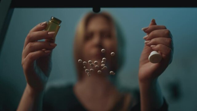 Depressed long haired woman plans suicide sitting at glass table with scattered small pills in semi-dark room at home close view low angle shot slow motion