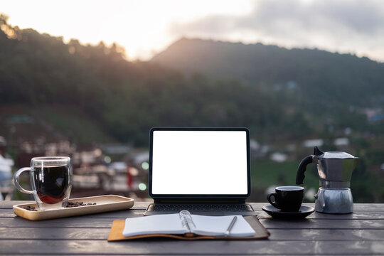 Morning cup of hot coffee and computer laptop on wooden table with mountain views in background.