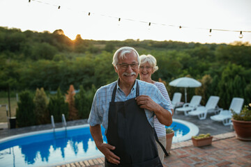 Senior woman standing behind her husband and tying him apron while standing near pool