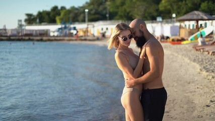 Young couple in love stand on the beach while on vacation and kiss passionately on the ocean shore. Spouses or lovers are resting at the resort.