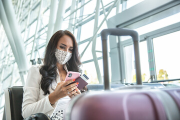 Woman in face mask checks her phone while waiting for an airplane in a hall, holding passport and a...
