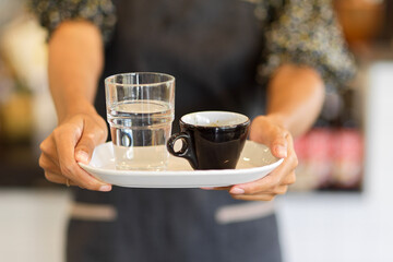Cropped image of black coffee mug with a glass of water on serving tray hold by female barista
