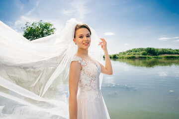 Fototapeta na wymiar a beautiful bride in a lace white dress with a long veil by the river 