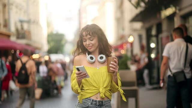 Young beautiful woman with dark curly hair walks around the city. The girl with one hand prints a message on the phone, and the other holds an eco cup of coffee