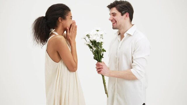 A happy man is giving flowers to his girlfriend standing isolated over white background in the studio
