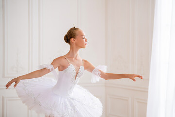 Fototapeta premium Portrait of young slender ballerina in a white tutu in elegant pose in large beautiful white hall.