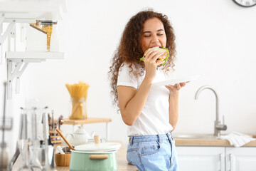 Young African-American woman eating tasty sandwich in kitchen