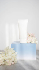 white cosmetic tube for cream on blue podium with flowers on white background with sunlight. Concept of gentle skin care. Vertical