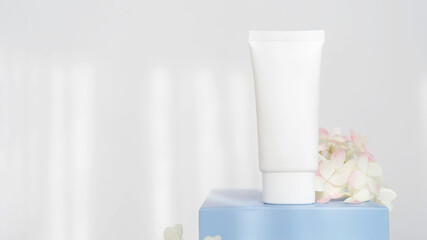 white cosmetic tube for cream on blue podium with flowers on white background with sunlight. Concept of gentle skin care. Copy space