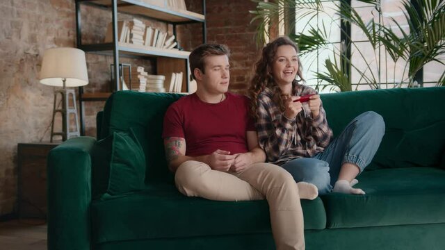 Young female sitting down on couch and grabbing controller from male gamer forcing man to take another gamepad and play videogame together on sofa at home