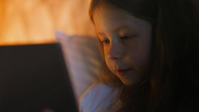 Adorable little girl talks looking at contemporary tablet computer on bed with pillows partially illuminated by night lamp light in shady room closeup slow motion