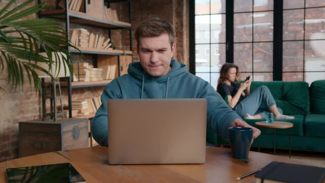 Pan right view of young man sipping energizing coffee from mug and flinching while sitting at table and working on remote project on laptop near girlfriend lounging on sofa