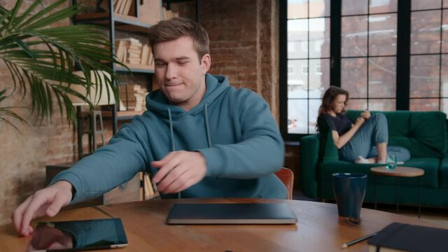 Pan left view of positive young guy closing laptop and starting to browse social media on tablet while sitting at table near girlfriend relaxing on couch at home