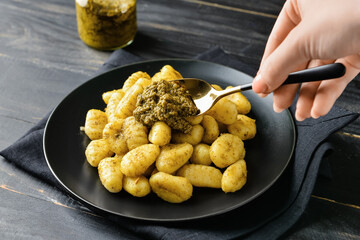 Woman adding pesto sauce into plate with tasty gnocchi on dark wooden background, closeup