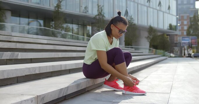 African woman in sportswear tying shoe laces sitting on stairs outdoors
