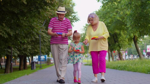 Senior grandmother grandfather walking with granddaughter playing squeezing anti-stress silicone toy simple dimple game in park. Family with pop it bubble squishy game with buttons. Stress reliever
