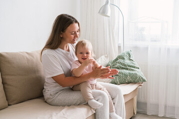 Horizontal of mum clapping and looking at blond child sitting on couch in living room. Mommy spending time with child at home. Concept of motherhood. Light background. Toddler looking away