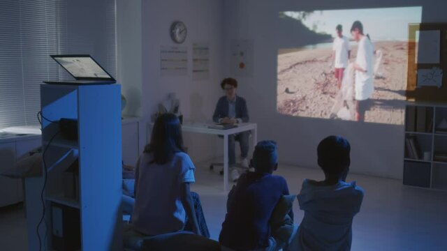 Handheld medium shot of female teacher showing educational movie about pollution to middle school children in dark classroom