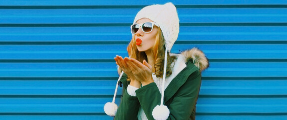 Winter portrait of beautiful young woman blowing her lips wearing a white hat with pom pom on blue background