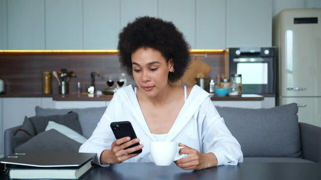 Confident portrait of a gorgeous young mixed race African American woman in casual clothes with a cup of coffee and a mobile phone in hands, resting sitting at the table on the home kitchen background