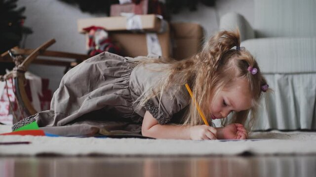 Close-up view 4k video footage of little cute baby girl sitting on floor in holiday cozy Christmas interior drawing pictures with pencil
