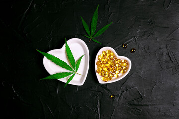Heart saucers with herbal cbd oil capsules and hemp leaves on black background.