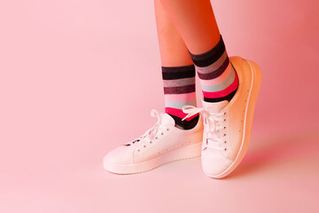 Fashion - white leather sneakers shoes, colourful striped socks and girl’s legs. Footwear on pastel pink background. Layout with free copy (text) space.