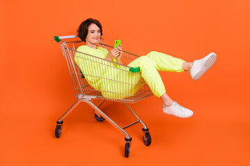 Portrait of attractive cheery funny focused girl sitting in shopping cart chatting blogging isolated over bright orange color background
