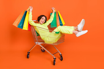 Portrait of attractive cheerful girl sitting in cart holding bags having fun holiday isolated over bright orange color background