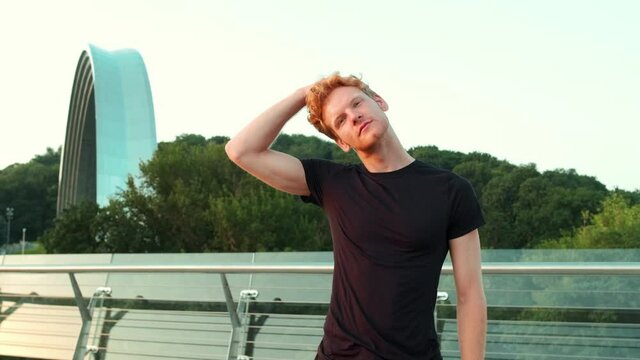 A handsome redhead man doing  warming-up exercises while standing on the bridge outside